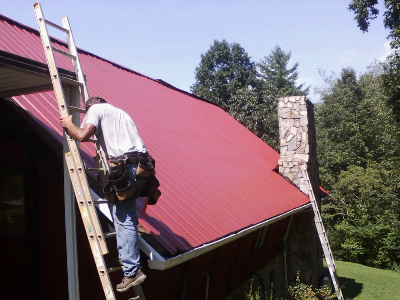 Roofer on a ladder beside a red metal roof and stone chimney — Soffit & Fascia Repair by Roofing Brothers in Petroleum